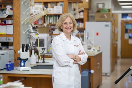 Frances Jensen, MD, chair of Neurology, wears a white coat and smiles with her arms folded while standing in a laboratory.